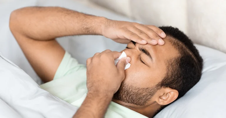 Man in bed holding tissue to blocked nose, struggling to breathe.