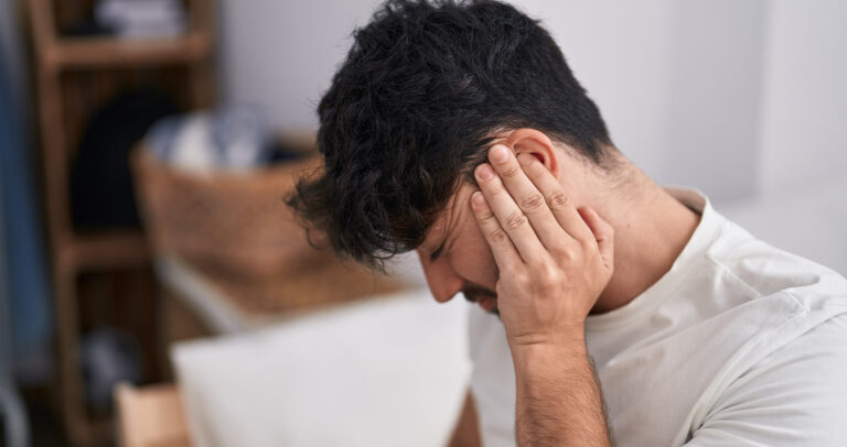 A man with dark curly hair and a white t-shirt presses his hand against his left ear with a pained facial expression.